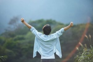 A person is standing with arms wide open to show success. The viewer sees the back of this person who faces a landscape with trees and mountains.